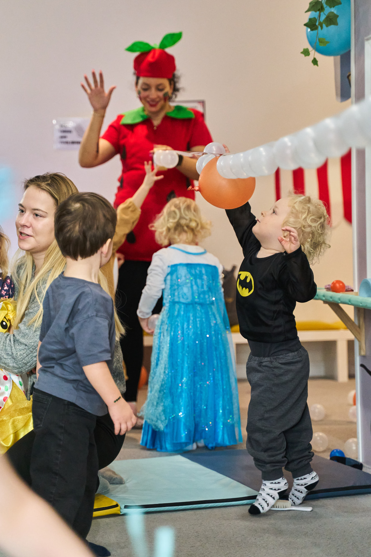 Dzieci i animatorka bawią się na przyjęciu urodzinowym w bawialni Mini Świat w Gdyni – chłopiec sięga po pomarańczowy balon, obok dziewczynka w błękitnej sukience księżniczki i uśmiechnięta animatorka w czerwonym stroju