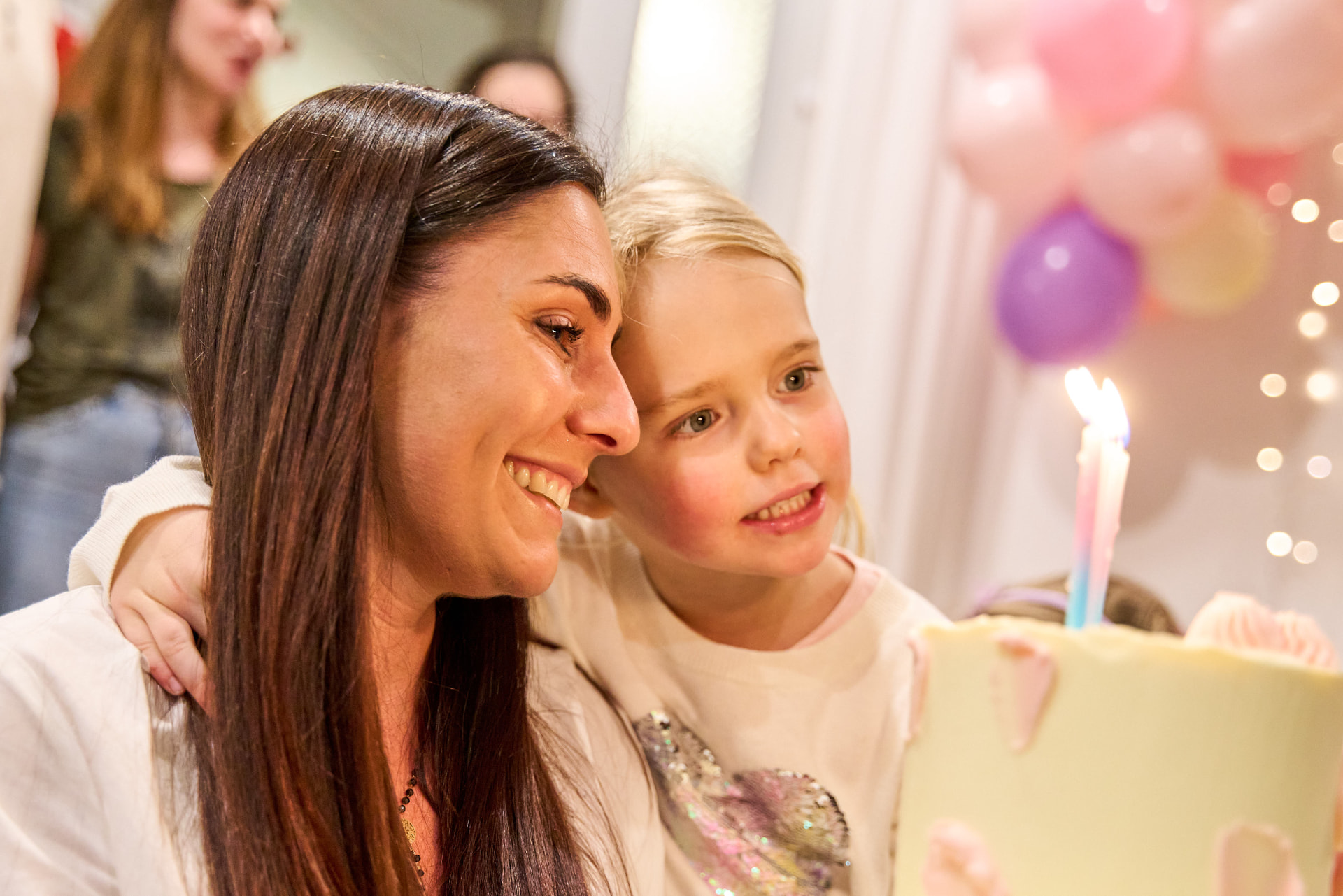 Kobieta i dziewczynka z uśmiechem patrzą na urodzinowy tort z zapalonymi świeczkami, w tle balony i girlandy świetlne w Mini Świecie w Gdyni
